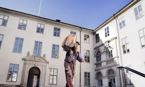 Man med skägg bär en stor träbit på axeln framför ett vitt, ståtligt medeltiden hus med många fönster.