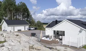 Två vita hus med svarta tak, en del av ett projekt för att bygga hus. En person sitter på en soffa utomhus, en barnvagn står bredvid.