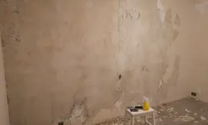 Förberedelser för att tapetsera på betong. Gamla tapeter skrapas bort från en sliten vägg. Verktyg och en sprayflaska står på en pall.