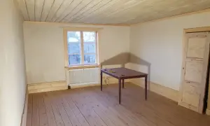 Ett rum med kalkputs inomhus, trägolv och en takbjälke. Ett fönster med utsikt över träd och ett bord med stolar.