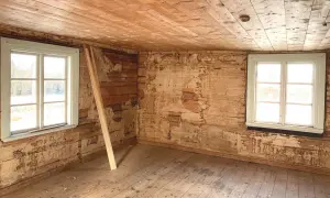 Ett rum i ett 1800-tals hus med synliga träväggar och ett trägolv. Två fönster släpper in ljus.