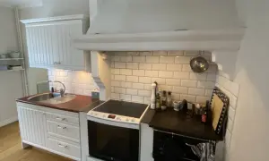 Vitt kök med en murad spiskåpa ovanför en vit spis och en diskho. Kakelvägg bakom spisen.