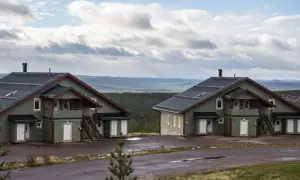 Två moderna knuttimrade stugor i grönt med mörka tak, belägna i ett kuperat landskap med skog och berg i bakgrunden.