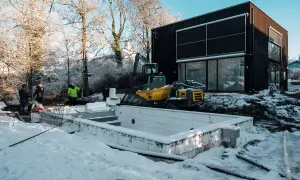 Snö täcker marken vid en pågående utbyggnad av ett hus. En grävmaskin och en tippkärra syns vid en nybyggd grund.