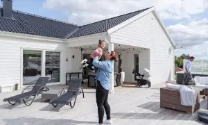 Kvinna lyfter barn på altanen framför ett av de färdiga husen. Barnvagn och utemöbler syns också.