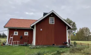 Rött torp med ny tillbyggnad och svensk flagga. Gräsklippare och grill står redo.