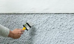 En hand målar på putsbruk med en pensel. Överdelen av väggen är slät och vit, medan den nedre delen har en grov, texturerad yta.