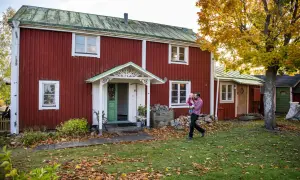 Man bär barn framför ett rött hus från 1700-talet med grönt tak. Höstlöv täcker gräsmattan.