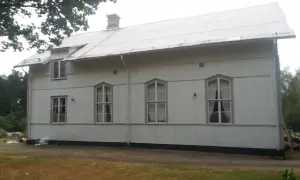 A white building with a corrugated metal roof and several windows, set against a backdrop of trees and a cloudy sky.