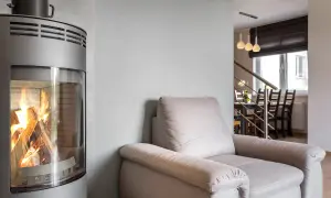 En modern kamin med brinnande ved. En bekväm fåtölj står bredvid, perfekt för en mysig kväll. Kanske dags att renovera kakelugn för en liknande känsla?