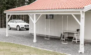 Vit bil parkerad vid en vit träfasad med röd tegeltak. En utbyggnad hus långsida med hängande gungor och ett bord.