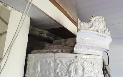 Detalj av en vit, ornamenterad kakelugn i lerbruk. En del av kaklet är skadat och visar den inre konstruktionen.