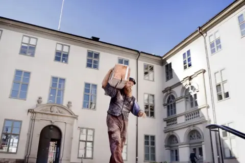 Man med skägg bär en stor träbit på axeln framför ett vitt, ståtligt medeltiden hus med många fönster.