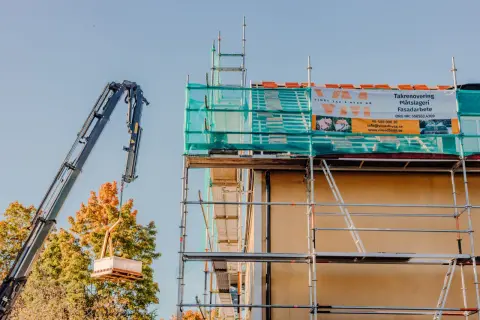 Kran lyfter material för att laga eternittak. Ställningar och byggnadsduk täcker huset.