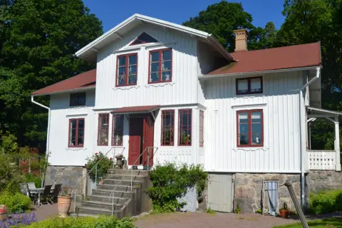 Vitt trähus med röda fönster och tak, en vacker **midja på gammalt hus** omgiven av grönskande träd.