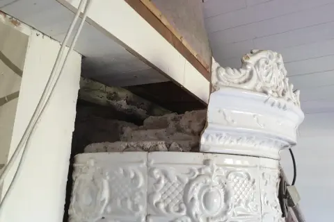 Detalj av en vit, ornamenterad kakelugn i lerbruk. En del av kaklet är skadat och visar den inre konstruktionen.