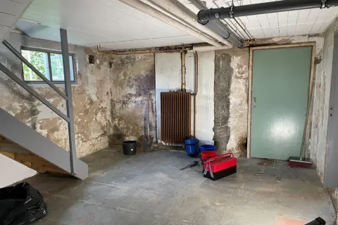 Källare med trappa, fönster, dörr, element och rör. Verktyg och hinkar står på golvet. Förberedelser för FTX ventilation och radonmätning.