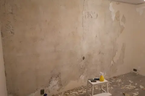 Förberedelser för att tapetsera på betong. Gamla tapeter skrapas bort från en sliten vägg. Verktyg och en sprayflaska står på en pall.