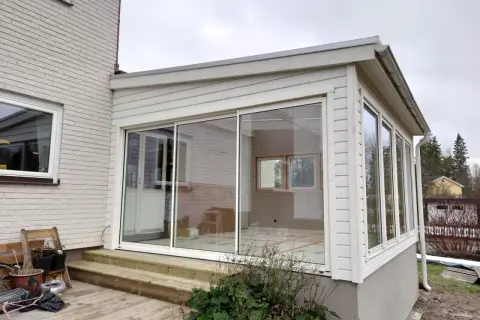 Ett modernt hus med glasveranda, med stora skjutdörrar i glas som släpper in mycket ljus. Trädäck och trappa leder upp till verandan.