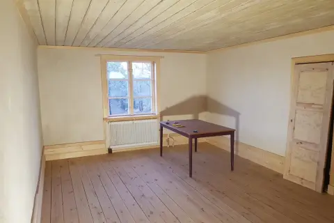 Ett rum med kalkputs inomhus, trägolv och en takbjälke. Ett fönster med utsikt över träd och ett bord med stolar.
