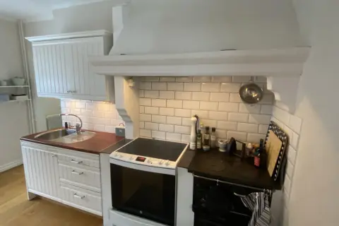 Vitt kök med en murad spiskåpa ovanför en vit spis och en diskho. Kakelvägg bakom spisen.
