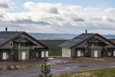 Två moderna knuttimrade stugor i grönt med mörka tak, belägna i ett kuperat landskap med skog och berg i bakgrunden.
