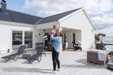 Kvinna lyfter barn på altanen framför ett av de färdiga husen. Barnvagn och utemöbler syns också.