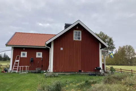 Rött torp med ny tillbyggnad och svensk flagga. Gräsklippare och grill står redo.