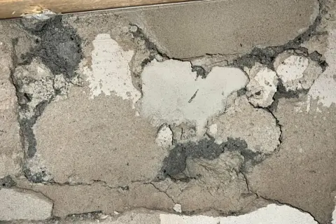 Skadad vägg med mörkgrå, smulig massa som kan vara blåbetong med asbest. Sprickor och avskalningar syns tydligt.