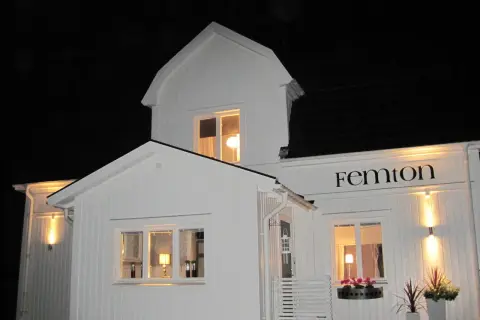 Vitt hus med svarta knutar, upplyst på kvällen. Namnet "Femton" syns ovanför ett fönster.