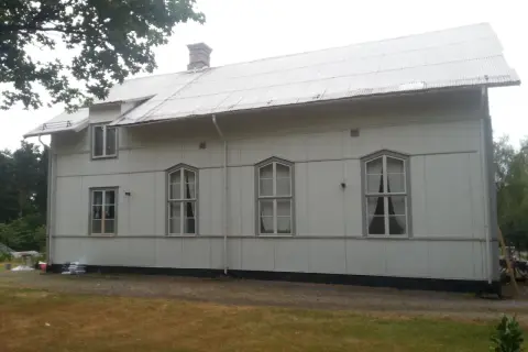 A white building with a corrugated metal roof and several windows, set against a backdrop of trees and a cloudy sky.