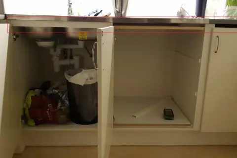 Under diskbänken pågår förberedelser för att installera diskmaskin i skåp. Syns rör, en papperskorg och diverse påsar.