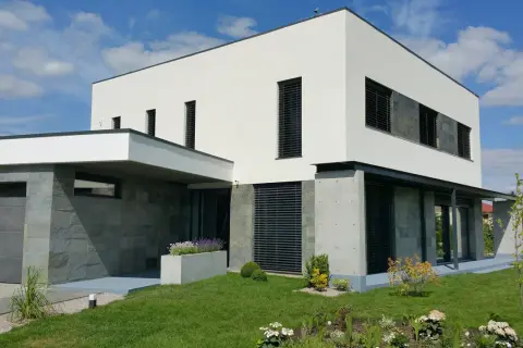 Modernt hus med vit, grå putsad fasad och stenbeklädda partier. Grönskande trädgård och garage.
