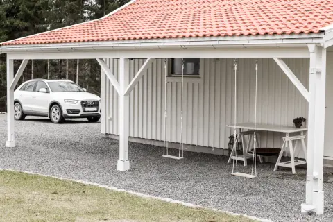 Vit bil parkerad vid en vit träfasad med röd tegeltak. En utbyggnad hus långsida med hängande gungor och ett bord.