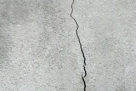 En spricka löper genom en grå, putsad fasad. Dags att laga sprickor i putsad fasad för att skydda huset.