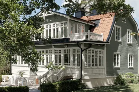 Ett charmigt grått hus med svarta knutar, omgivet av grönska. Ett vackert hus med många fönster och en veranda.