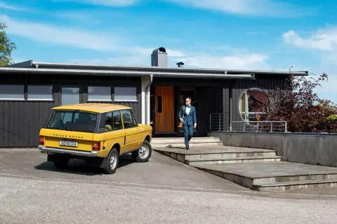 Man i kostym går mot ett funkishus från 60-talet med en gul Range Rover parkerad utanför.