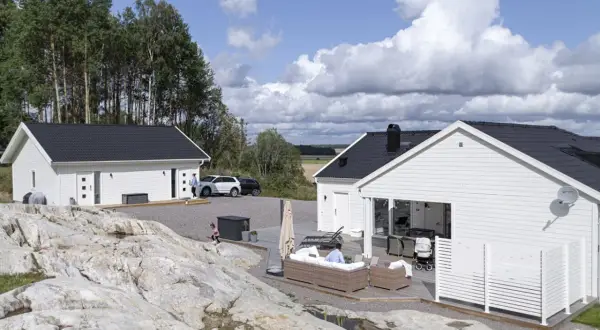 Två vita hus med svarta tak, en del av ett projekt för att bygga hus. En person sitter på en soffa utomhus, en barnvagn står bredvid.