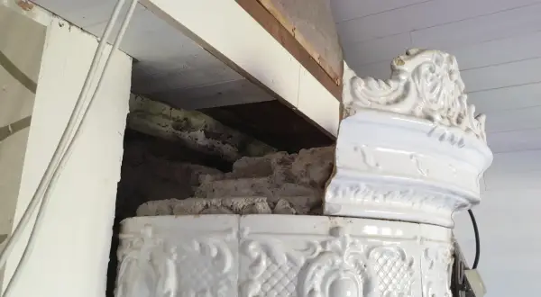 Detalj av en vit, ornamenterad kakelugn i lerbruk. En del av kaklet är skadat och visar den inre konstruktionen.