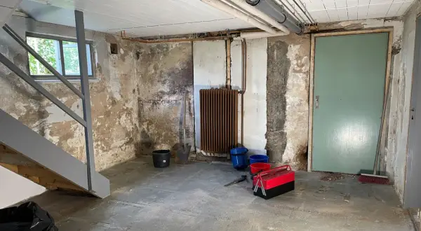 Källare med trappa, fönster, dörr, element och rör. Verktyg och hinkar står på golvet. Förberedelser för FTX ventilation och radonmätning.