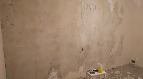 Förberedelser för att tapetsera på betong. Gamla tapeter skrapas bort från en sliten vägg. Verktyg och en sprayflaska står på en pall.