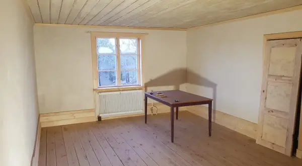 Ett rum med kalkputs inomhus, trägolv och en takbjälke. Ett fönster med utsikt över träd och ett bord med stolar.