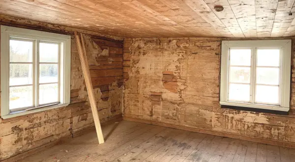 Ett rum i ett 1800-tals hus med synliga träväggar och ett trägolv. Två fönster släpper in ljus.