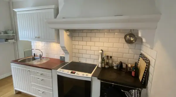 Vitt kök med en murad spiskåpa ovanför en vit spis och en diskho. Kakelvägg bakom spisen.