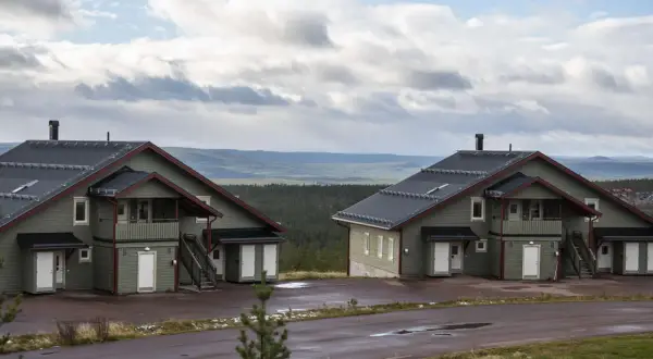 Två moderna knuttimrade stugor i grönt med mörka tak, belägna i ett kuperat landskap med skog och berg i bakgrunden.