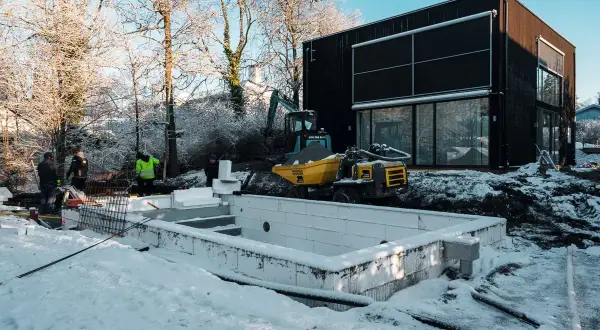 Snö täcker marken vid en pågående utbyggnad av ett hus. En grävmaskin och en tippkärra syns vid en nybyggd grund.