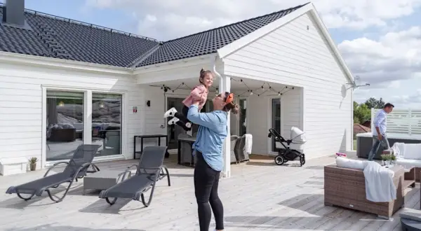 Kvinna lyfter barn på altanen framför ett av de färdiga husen. Barnvagn och utemöbler syns också.