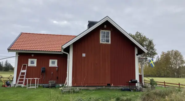 Rött torp med ny tillbyggnad och svensk flagga. Gräsklippare och grill står redo.