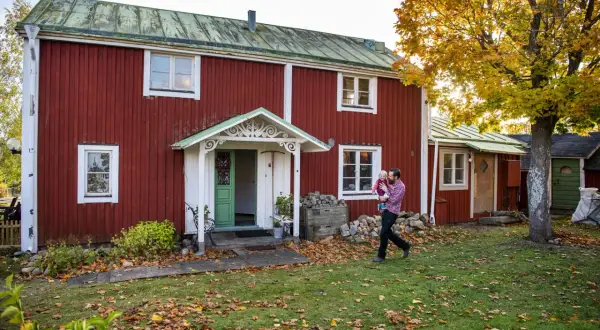 Man bär barn framför ett rött hus från 1700-talet med grönt tak. Höstlöv täcker gräsmattan.