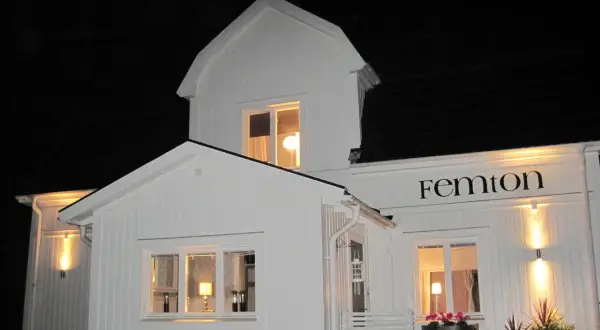Vitt hus med svarta knutar, upplyst på kvällen. Namnet "Femton" syns ovanför ett fönster.